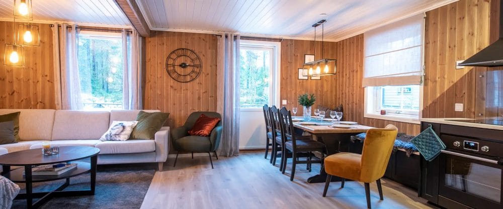 Bright living room with wood panelling, white sofa, dining table with six chairs, large windows and modern kitchen area.