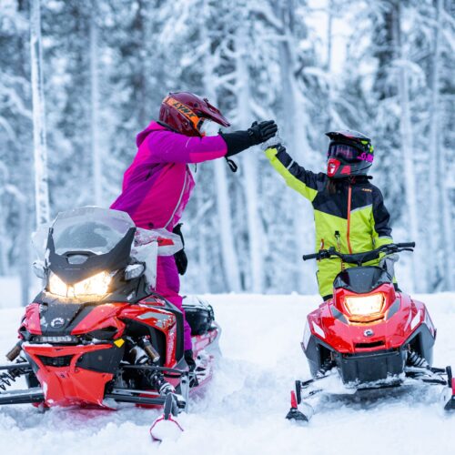 Two people on red snowmobiles high-five in snow-covered forest during winter tour.