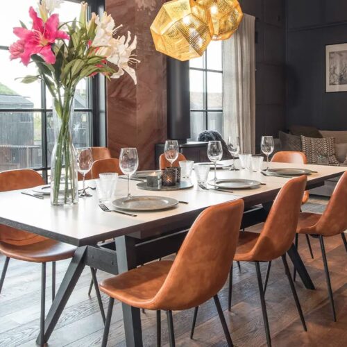 Dark living room with large windows, dining table set with six chairs, flowers in vase, sofa corner and modern lamps.