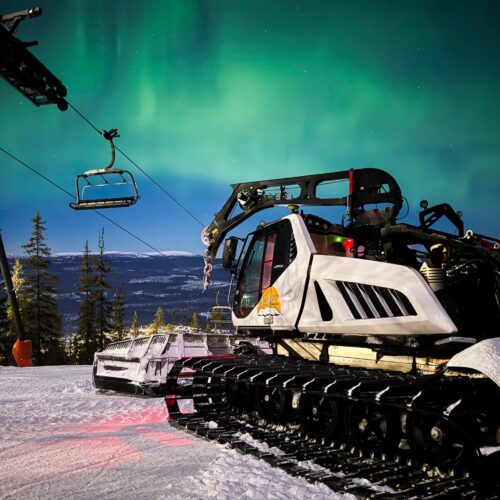 Snow groomer standing by ski lift on snow-covered slope under northern lights and starry sky in mountain environment.