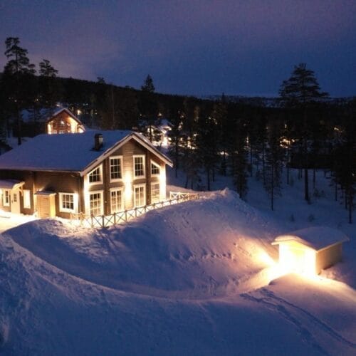 Oplyst bjerghytte i snedækket landskab om aftenen med omgivende skov og hytter.