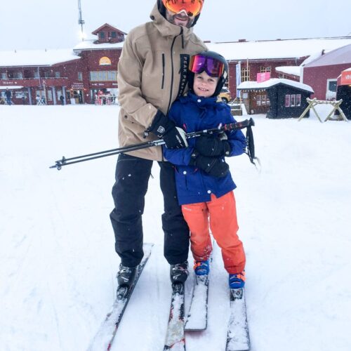 Two people standing on skis in a snowy ski area in front of red buildings with poles in their hands.