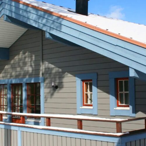Grey wooden cottage with blue details, snow-covered roof and surrounding pine forest in sunny weather.