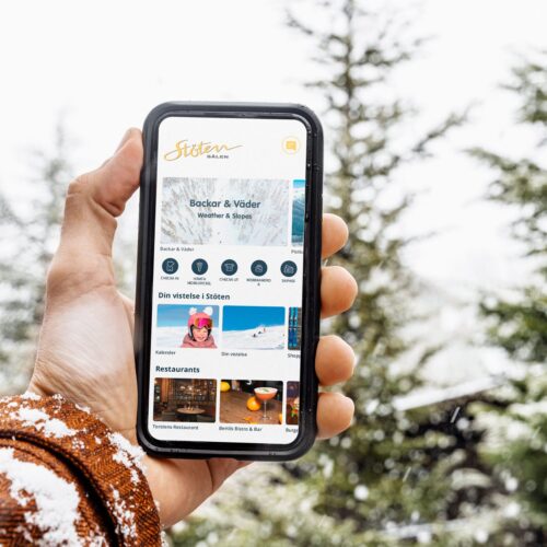 Hand håller smartphone med Stöten Sälen-app som visar backar, väder och restauranger, snöiga träd i bakgrunden.