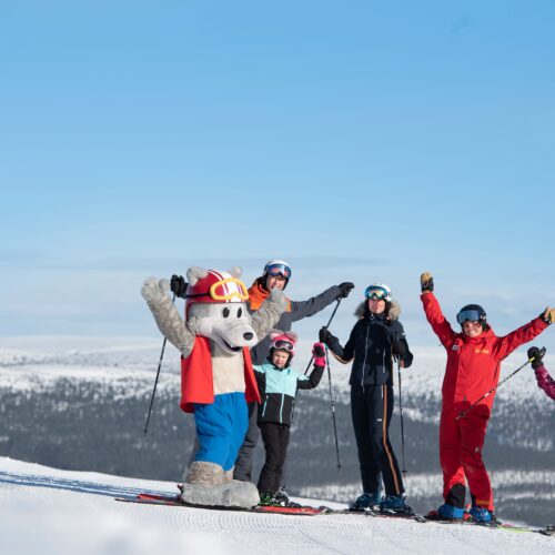 En gruppe skiløbere og maskotten Vargy står på et snedækket bjerg med stave under en klar blå himmel.