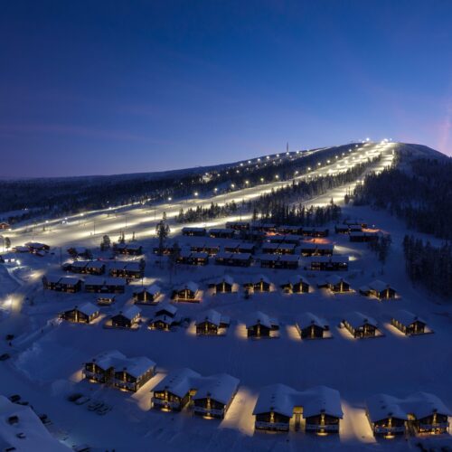 Snow-covered ski resort in evening light with illuminated ski slopes, chalets and roads, surrounded by dark forests and mountains under blue sky