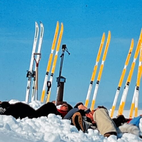 Several people are lying in the snow, sunbathing with cross-country skis against a clear blue sky.