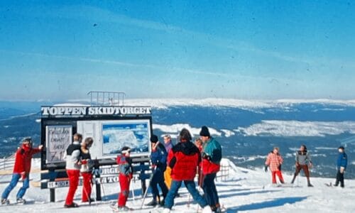 Skiløbere samledes ved informationstavlen på bjergtoppen med udsigt til snedækkede bjerge og blå himmel.