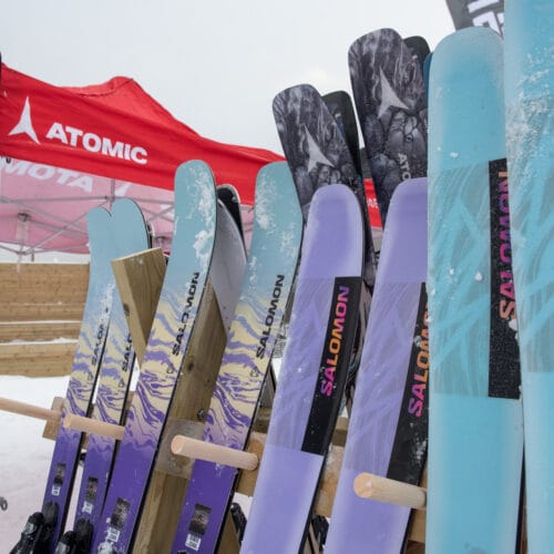 Several Salomon skis in different colours are lined up under a red Atomic canvas at the ski area.