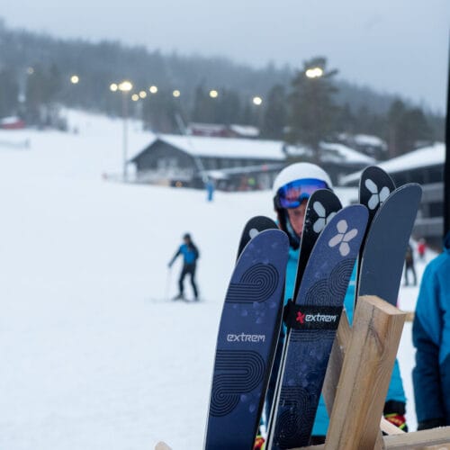 Ski med Extrem-logoet er parkeret på skibakken med skiløbere og hytter i baggrunden.