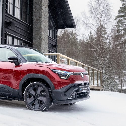 Röd SUV parkerad på snötäckt uppfart vid mörkt trähus med skog i vinterlandskap.