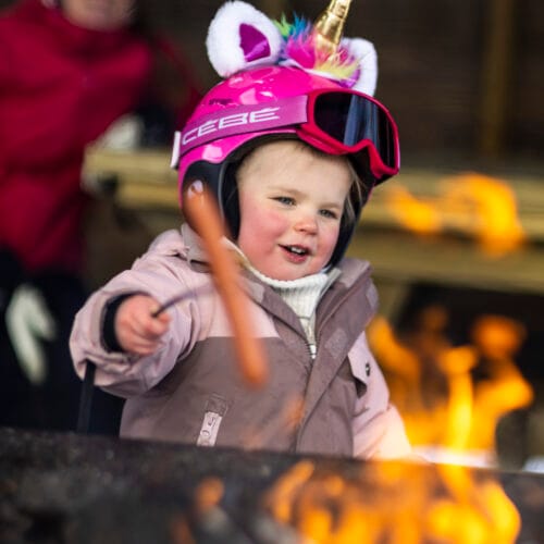 Børn med lyserøde enhjørningshjelme griller pølser over åben ild med skitøj i baggrunden.