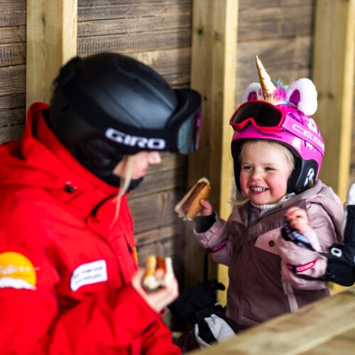 Två personer i skidkläder äter korv i träbod, barn med rosa enhörningshjälm