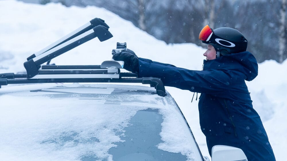 En person står ved en bil og spænder ski på taget i et vinterlandskab.