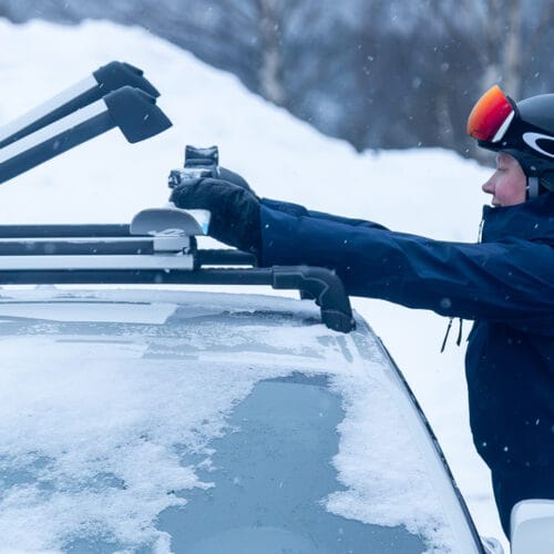 En person står ved en bil og spænder ski på taget i et vinterlandskab.