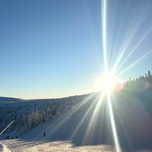 Snow-covered ski slope at sunrise with piste machine tracks and forested mountains in the background.