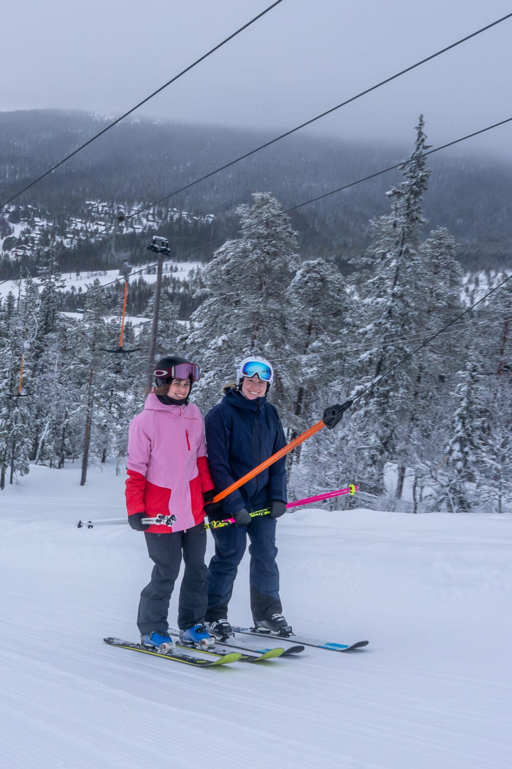 Two skiers on a snowy slope with lift, winter landscape and fir trees.