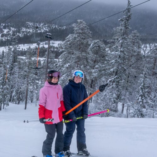 Two skiers on a snowy slope with lift, winter landscape and fir trees.