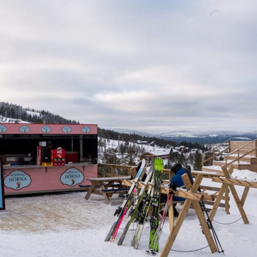 Ski racks and wooden benches in front of a pink catering kiosk in a snow-covered mountain environment with a wide view.