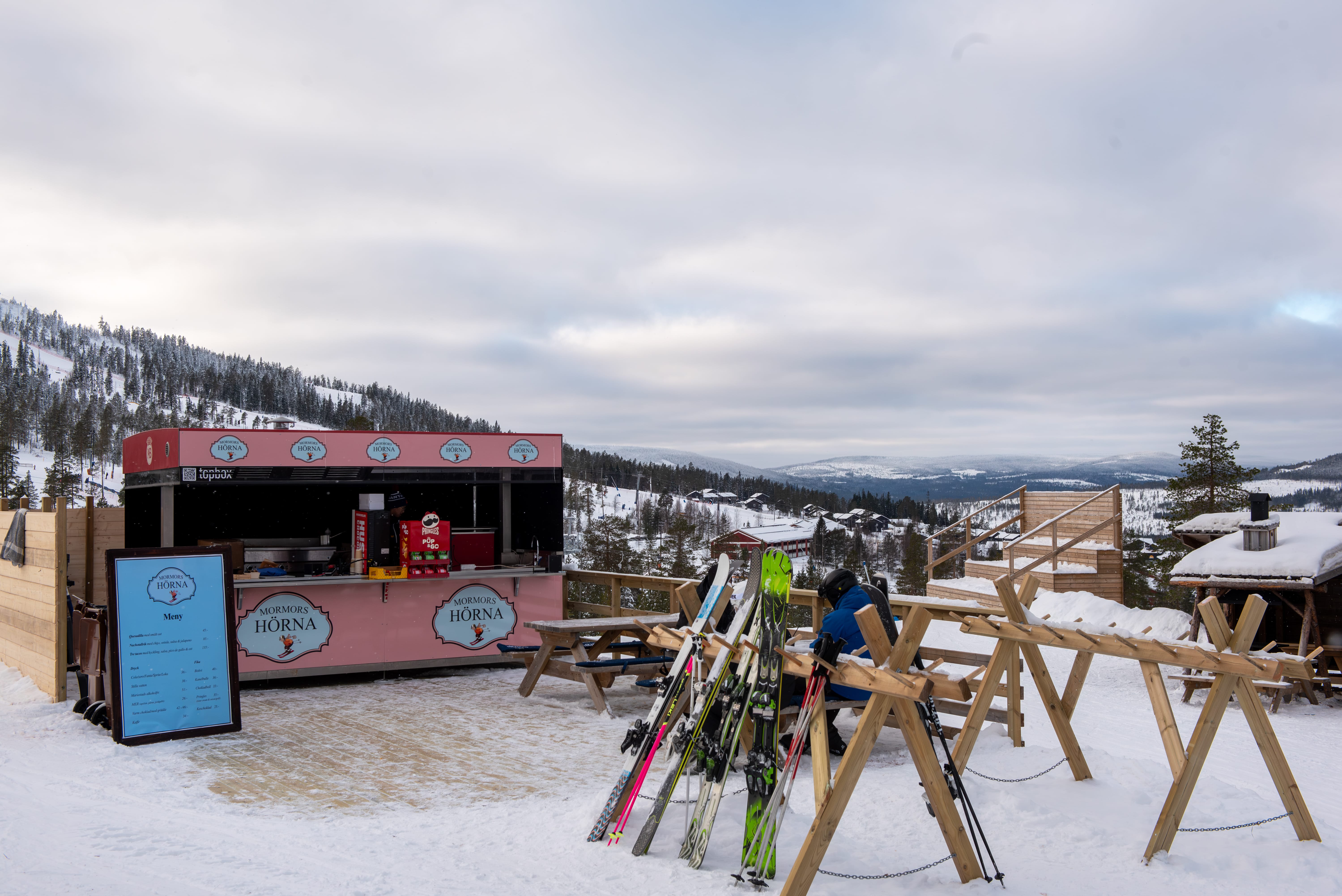 Skidställ och träbänkar framför rosa serveringskiosk i snötäckt fjällmiljö med vid utsikt.