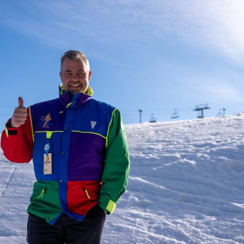 Happy person gives a thumbs up wearing a colourful jacket in front of a sunny ski slope