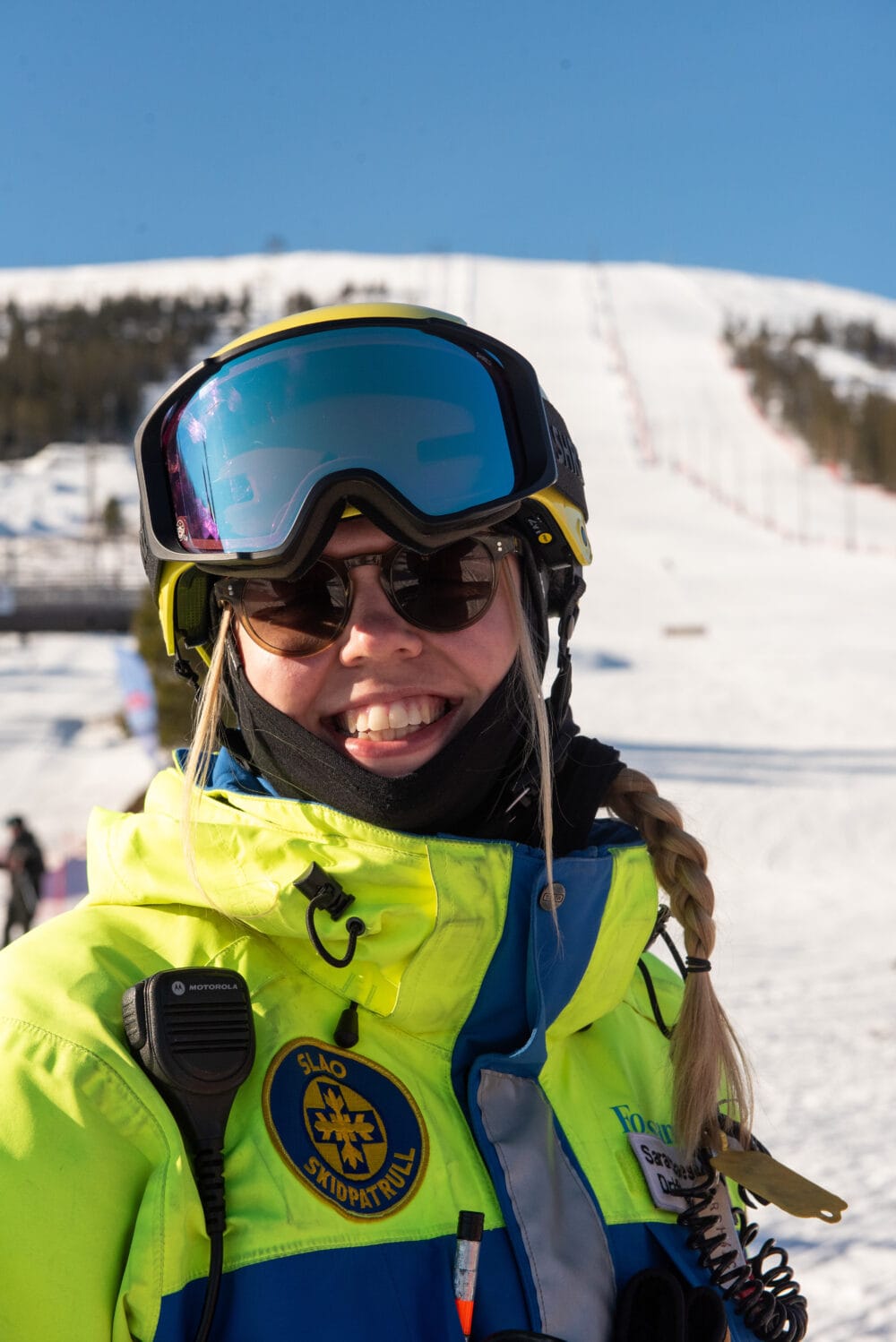 Person wearing a yellow-blue ski suit and ski helmet with ski goggles smiles at the camera.