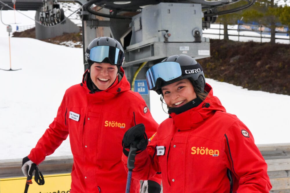 Two ski instructors wearing red ski suits and smiling at the camera while waiting for the lift.