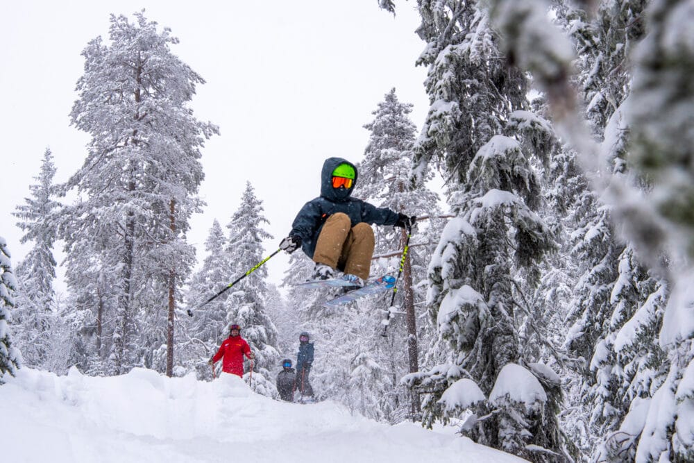 Children skiing in a snow-covered forest with a ski instructor looking on.