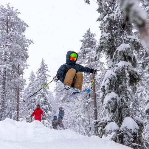 Barn hoppar på skidor i en snöklädd skog med en skidlärare som tittar på.