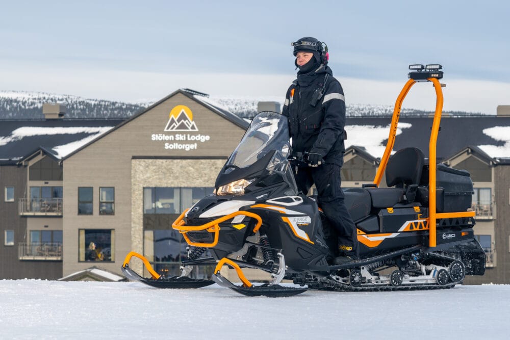 Mechanics sitting on a snowmobile in front of a SkiLodge in a mountainous landscape.