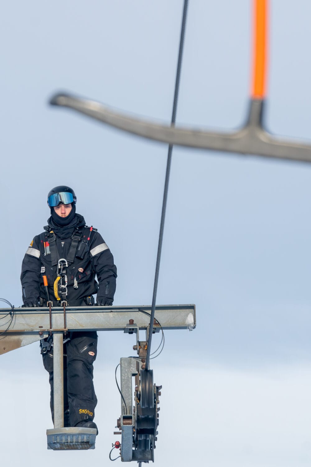 A mechanic stands at the top of a lift mast wearing a black uniform.