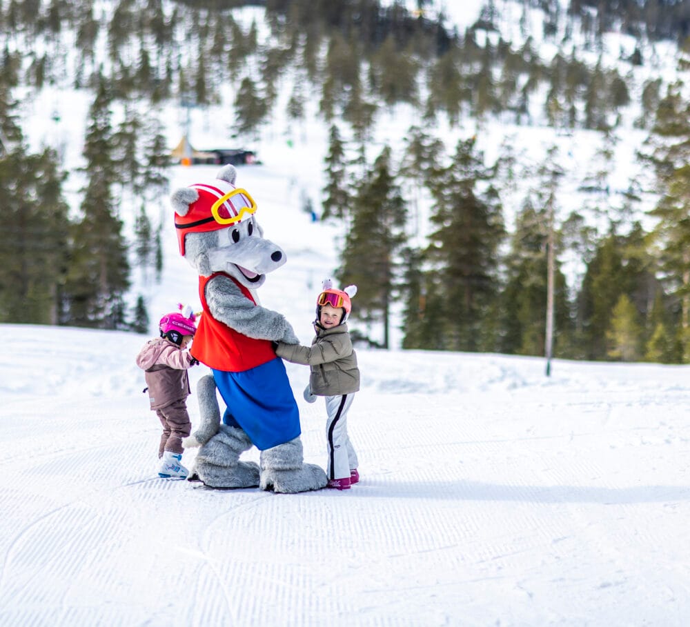 Maskoten Vargy går i en skidbacke tillsammans med två barn.