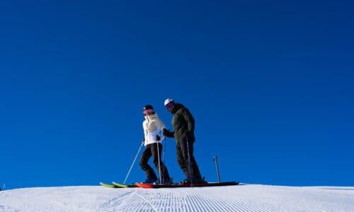 Two people are standing on a ski slope with blue skies and freshly groomed snow.