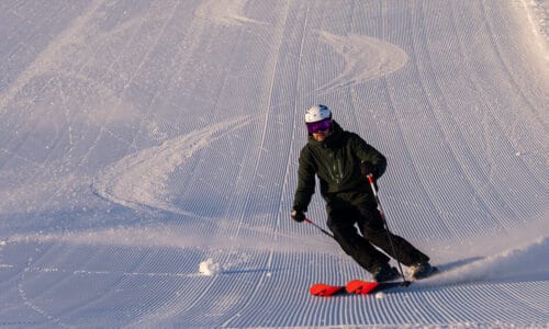 A skier in a dark jacket turns down a freshly groomed piste in sunny winter light.