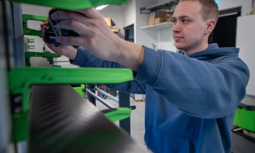 Person justerar maskinkomponent på grön industriell utrustning i verkstadsmiljö med verktyg och hyllsystem i bakgrunden.