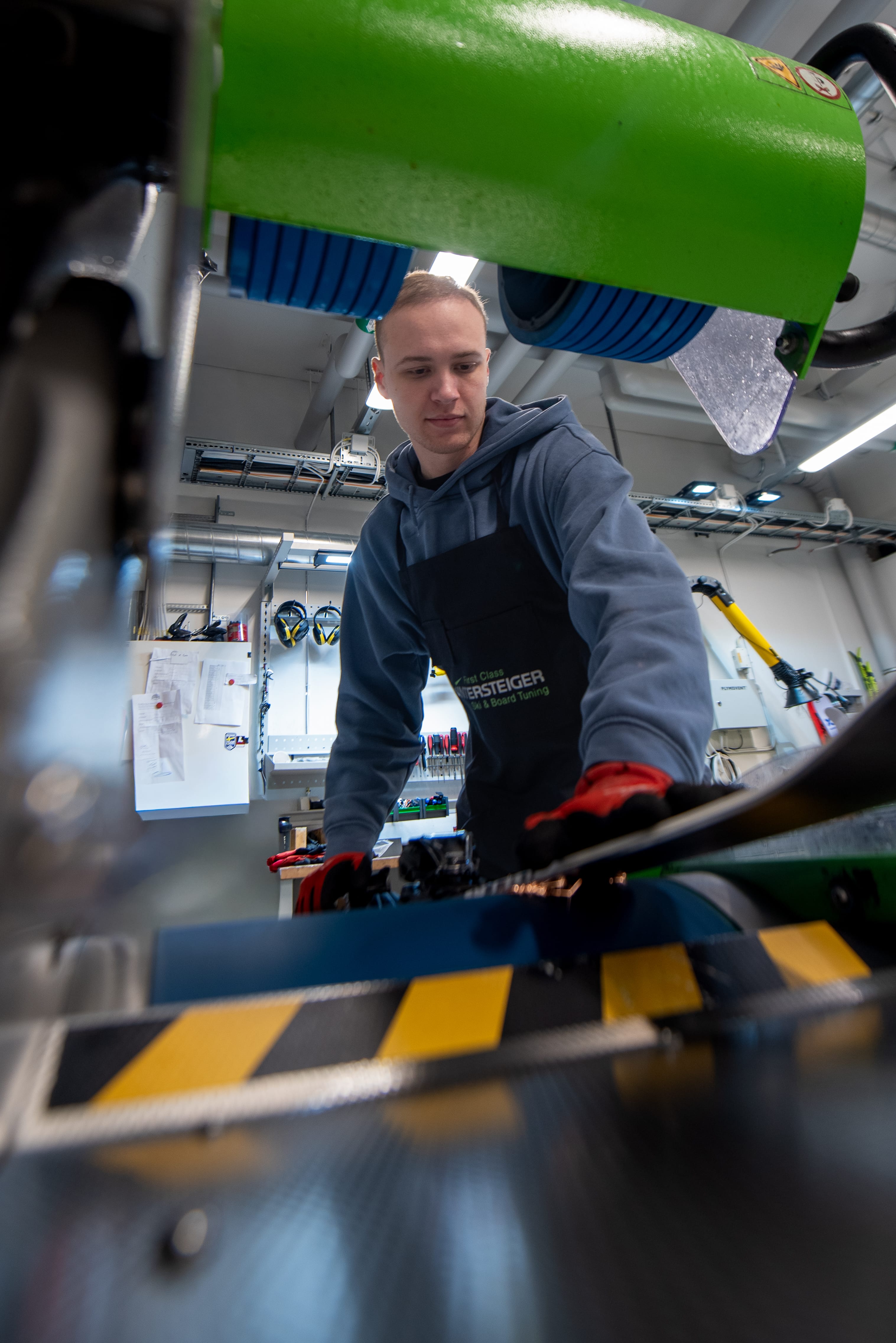 Person justerar maskin på industriell arbetsstation med skyddshandskar och gröna maskindelar i verkstad.
