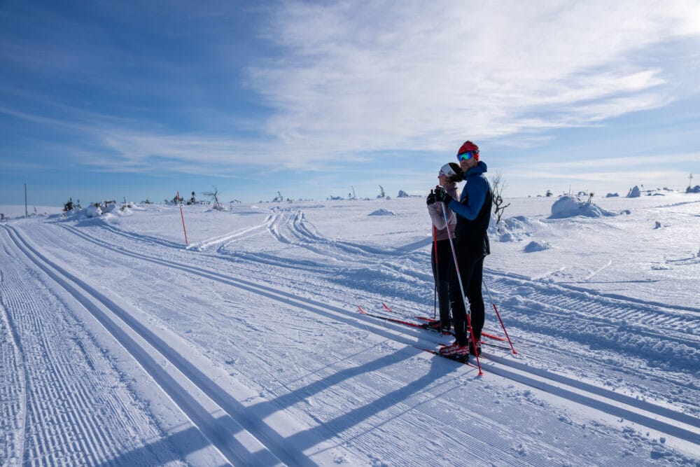 Två personer står på längdskidor i preparerat spår öppet snölandskap under blå himmel.